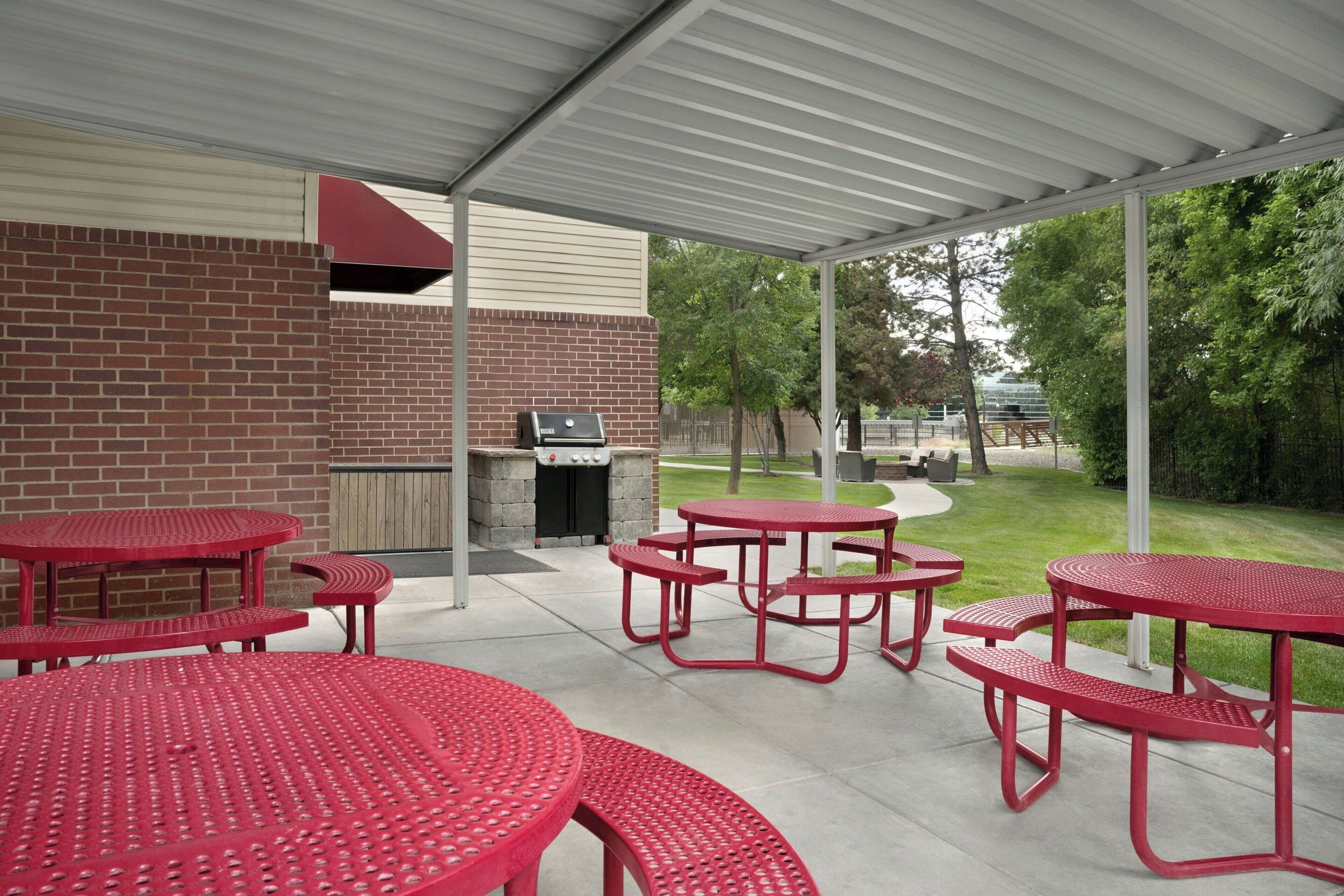 A covered patio area with red metal tables and benches, a barbecue grill, and a brick wall.