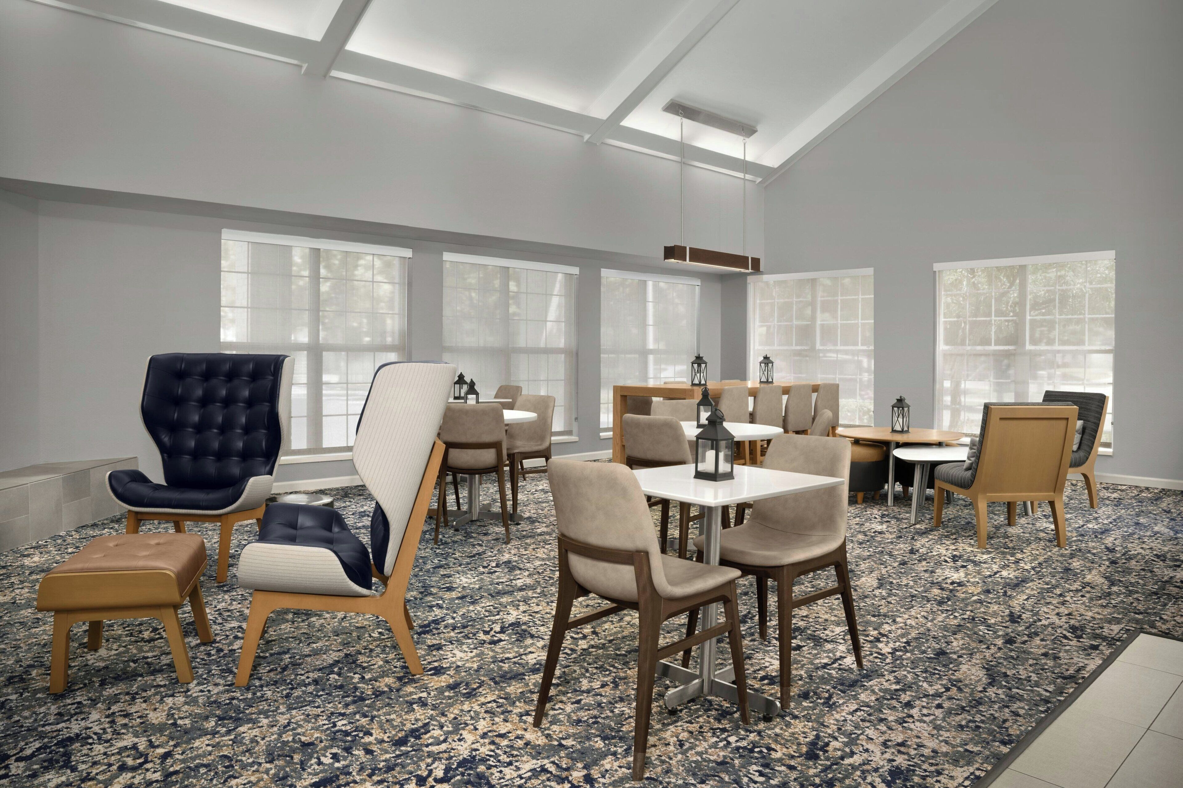 A spacious room with a high ceiling, large windows, and a mix of seating arrangements including chairs and tables.