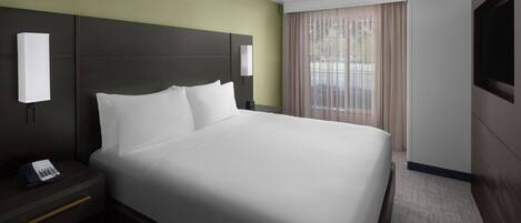 A hotel room with a large bed, a bedside table with a phone, a wall-mounted TV, and a window with curtains.