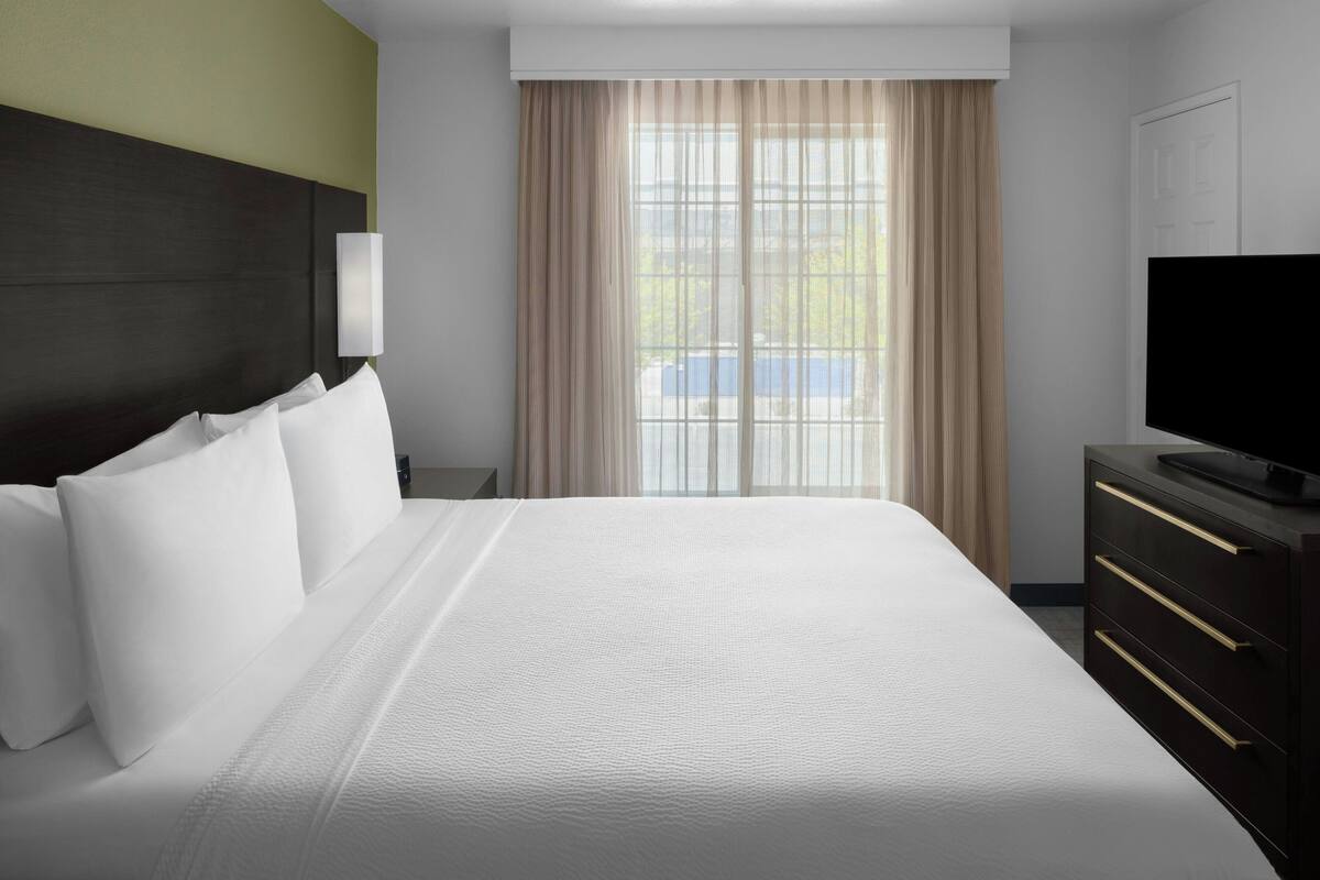 Una habitación de hotel con una cama grande, un televisor, un aparador y una ventana con cortinas.