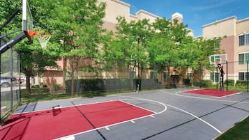 An outdoor basketball court with a red surface, surrounded by a chain-link fence and trees.