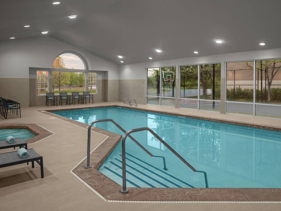 Una piscina interior con paredes de vidrio transparente, un área de descanso separada con sillones, y un aro de baloncesto visible a través de las ventanas.