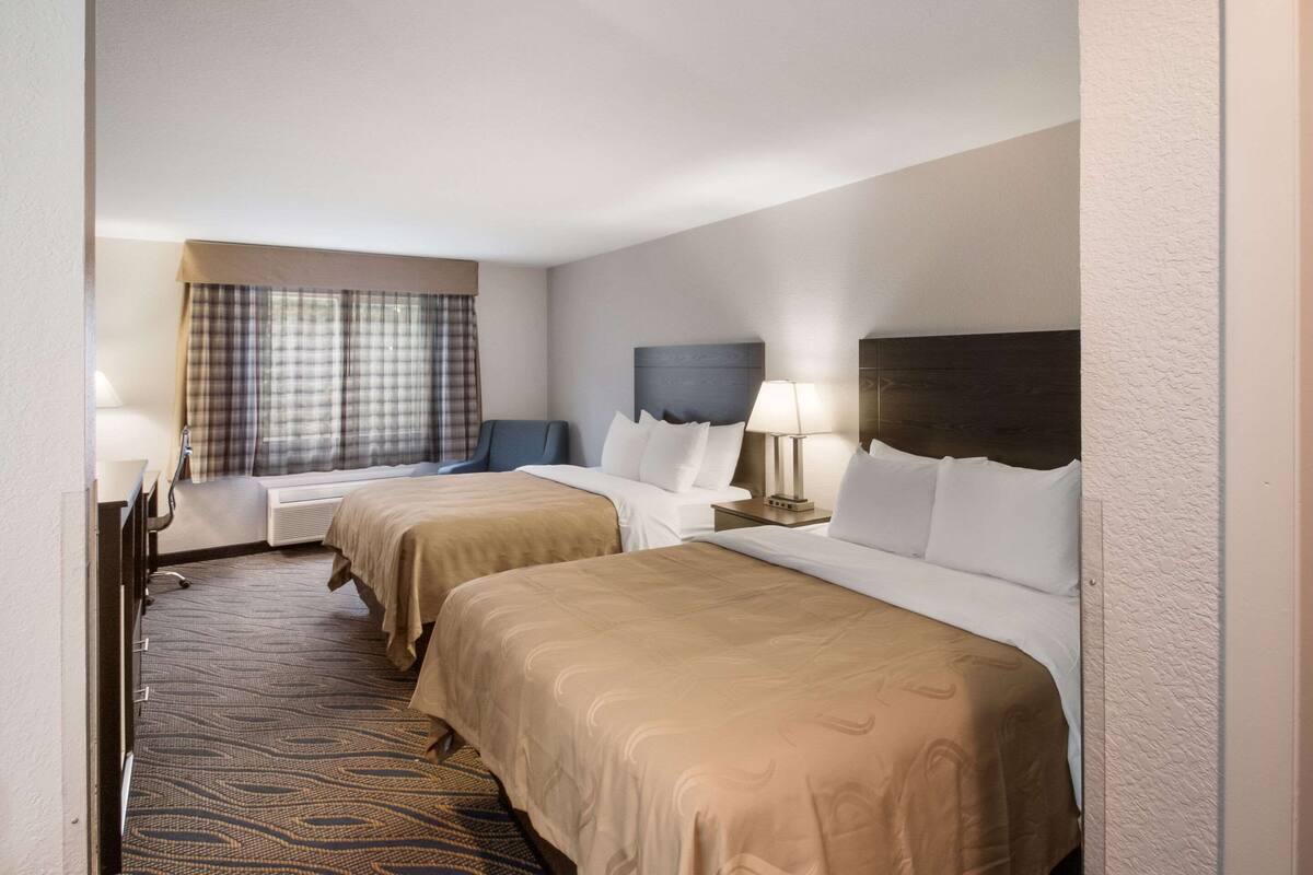 Ein Hotelzimmer mit zwei Betten, einem Schreibtisch, einem Stuhl, einem Fenster mit Vorhängen und einem Kopfteil.