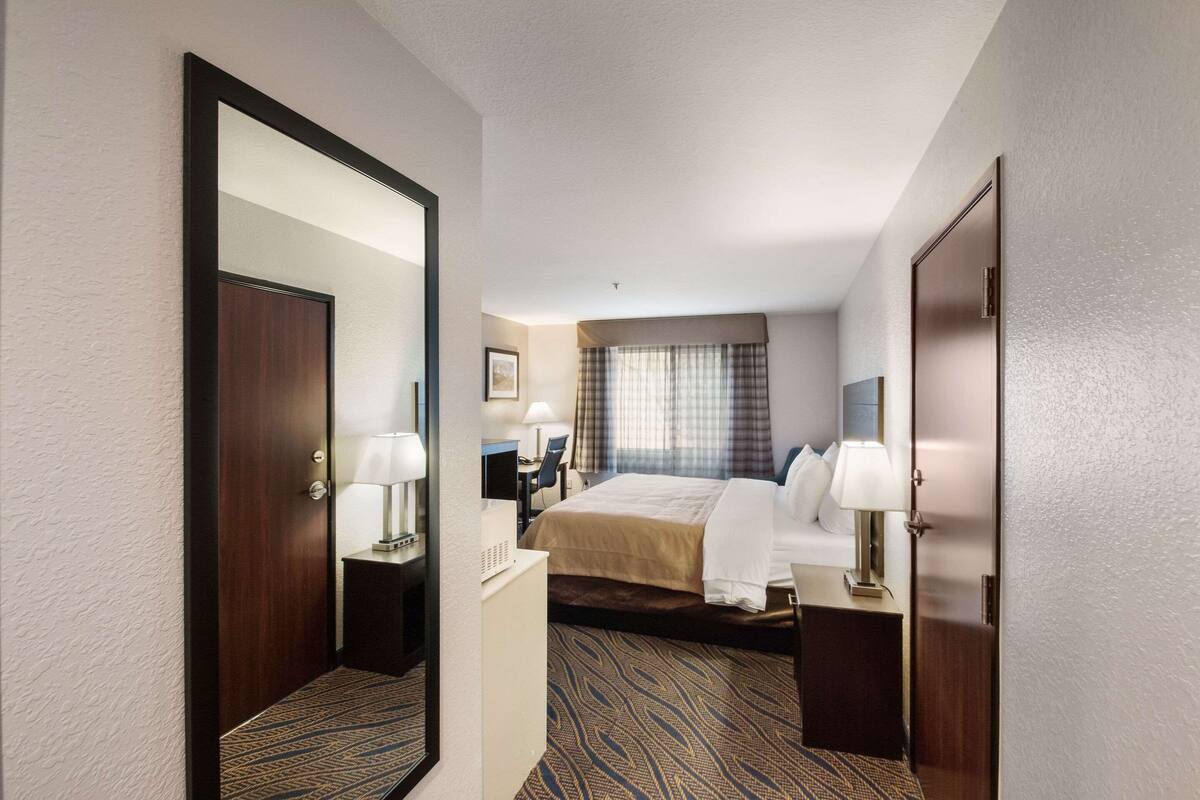 Ein Hotelzimmer mit einem großen Bett, einem Schreibtisch mit Stuhl, einem Spiegel und einem Fenster mit Vorhängen.