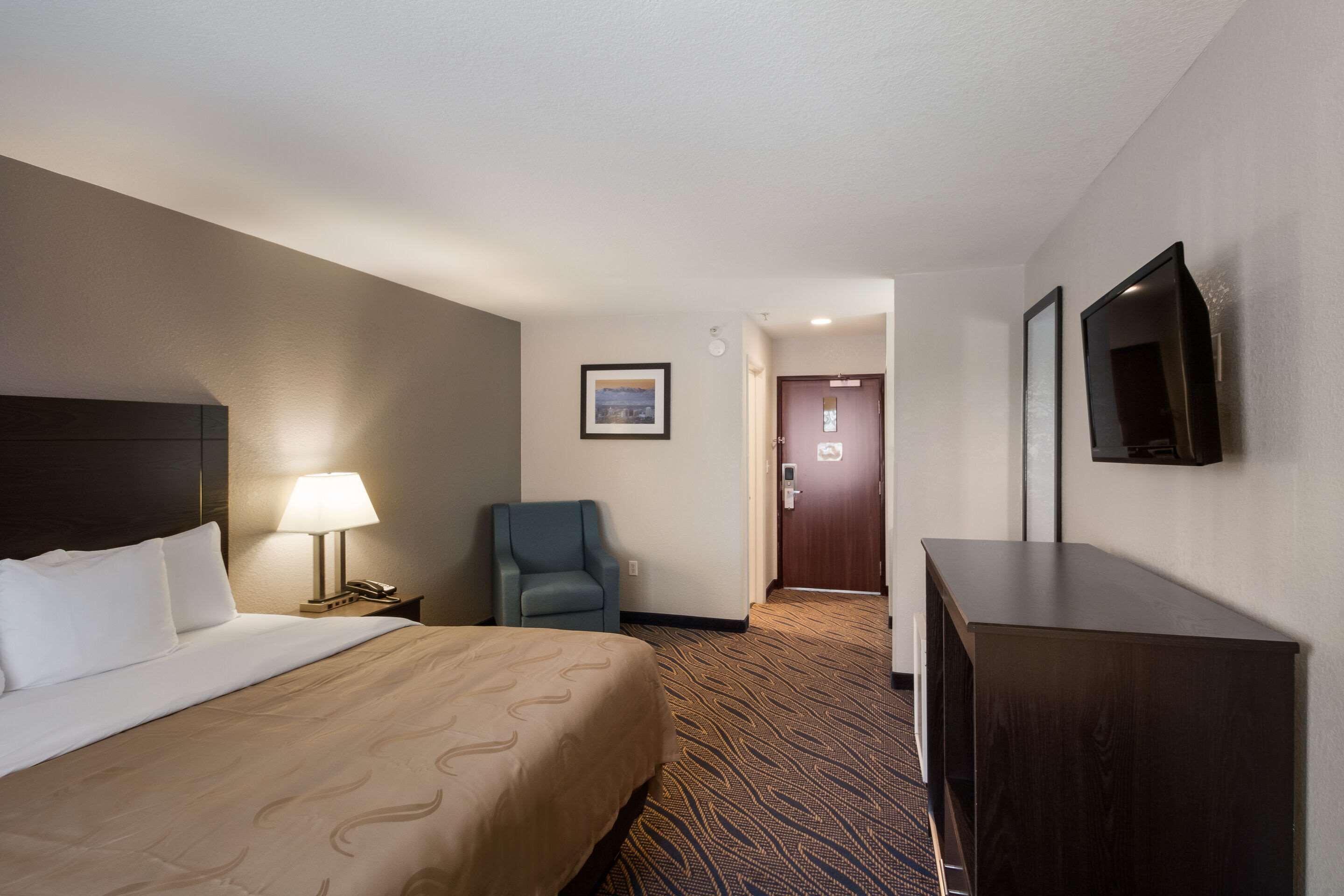Quarto de hotel com uma cama, uma mesa com luminária, uma cadeira, uma televisão e uma porta que dá para outro cômodo.