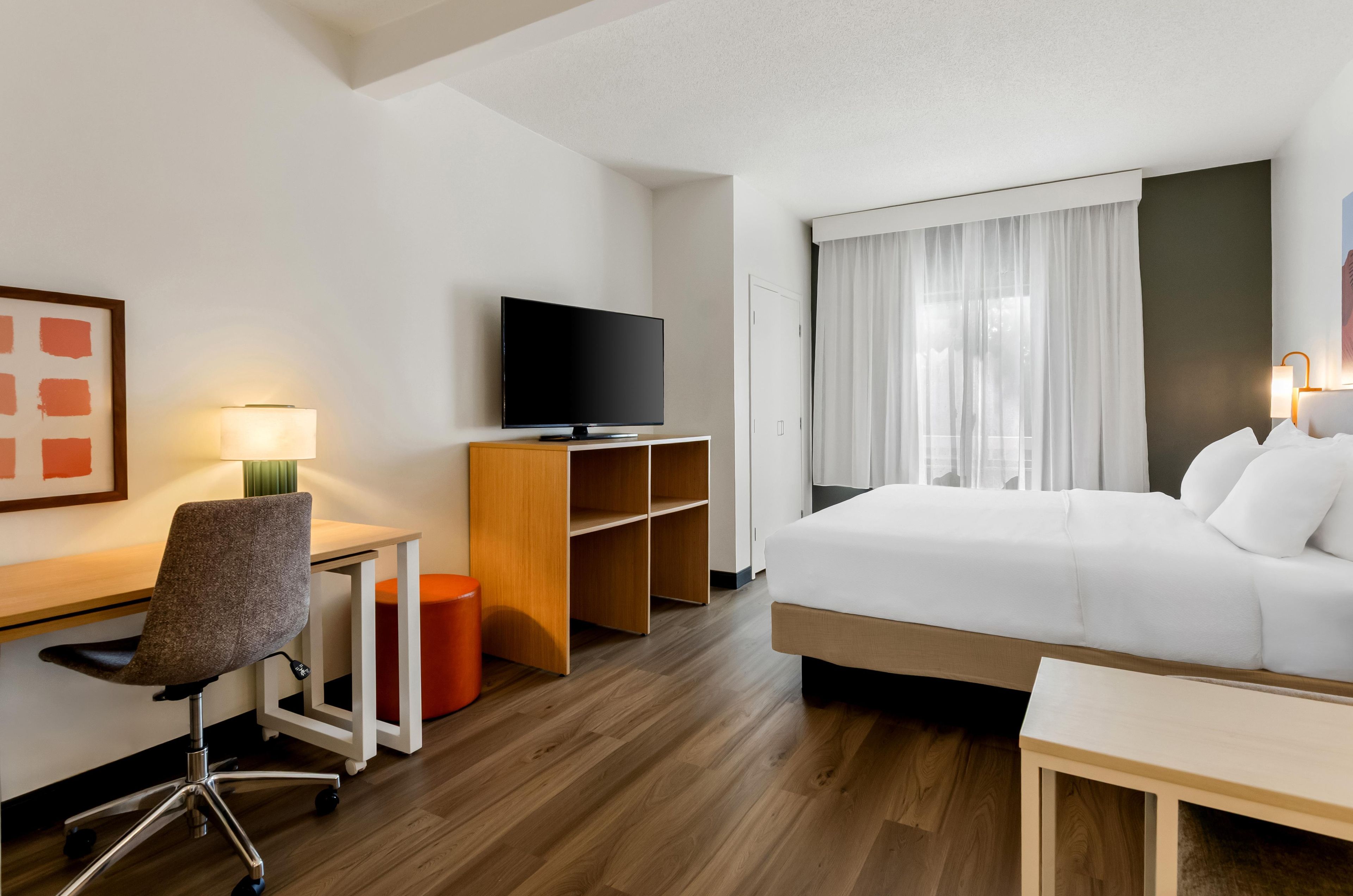 Una camera d'albergo con un letto, una scrivania, una sedia, una televisione e una finestra con tende.