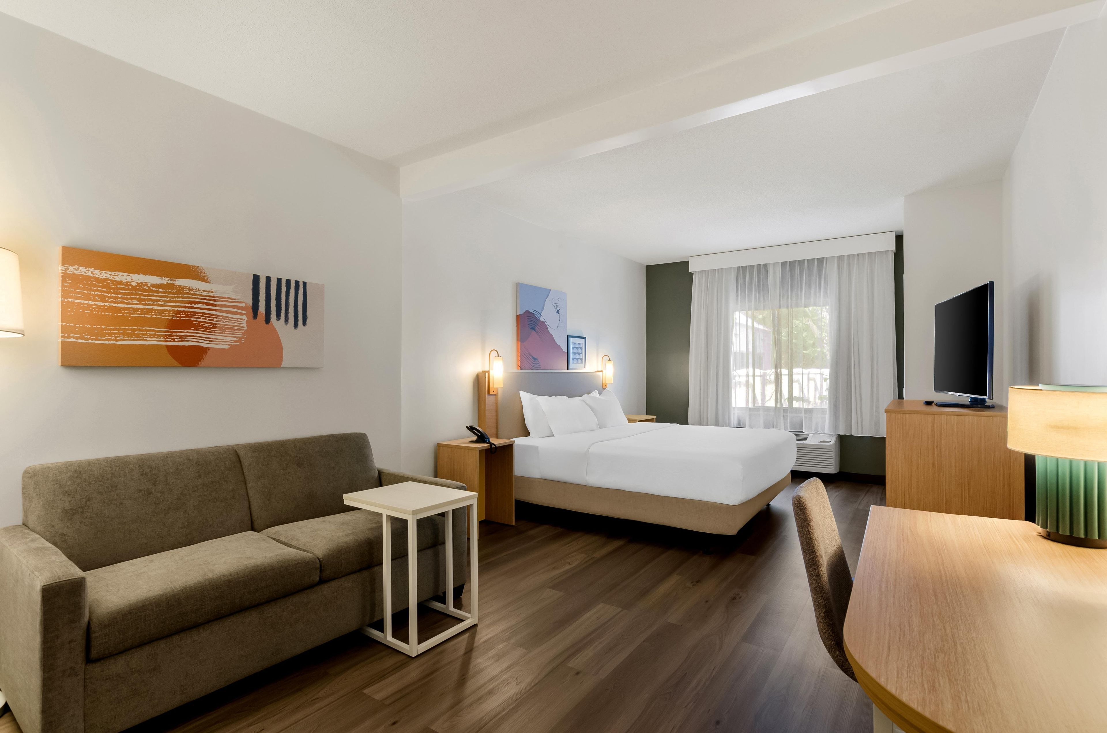 A hotel room with a bed, a sofa, a small table, a TV, and artwork on the walls.