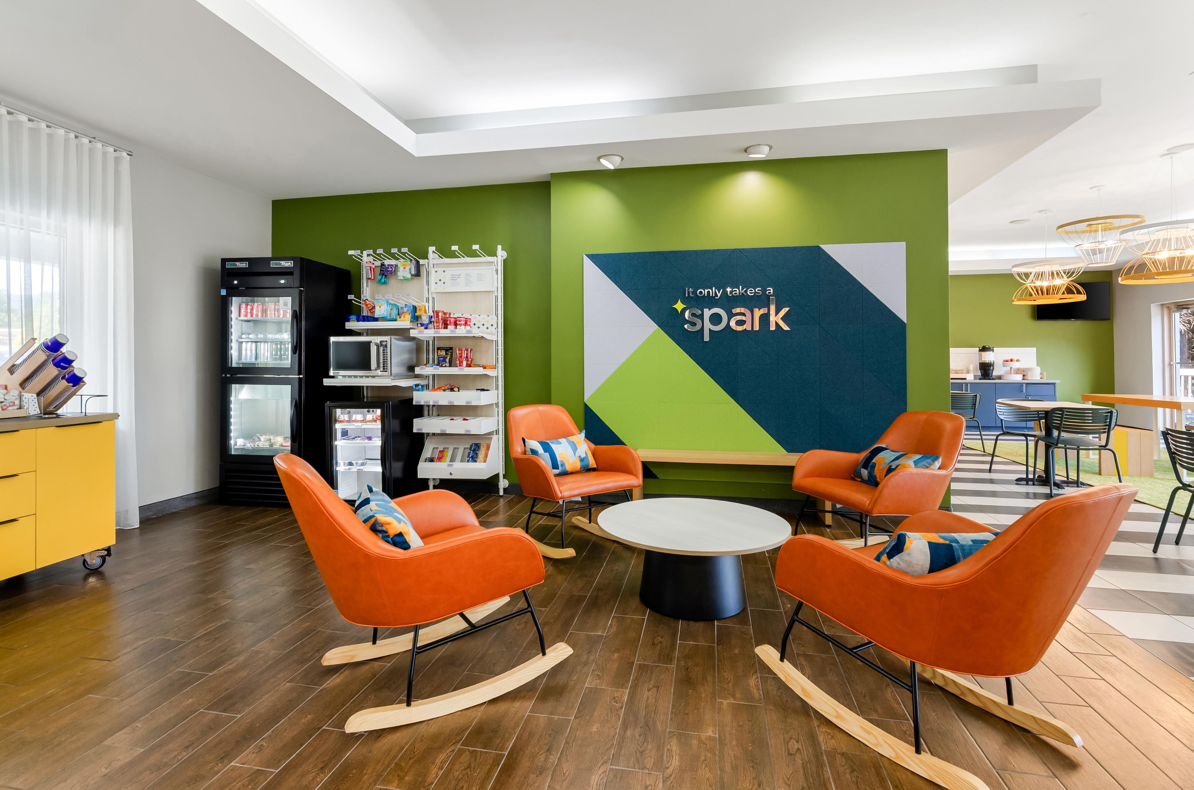 Un espace de détente moderne avec des fauteuils à bascule orange, une table ronde et un mur vert orné d’un panneau indiquant le mot « Spark ».