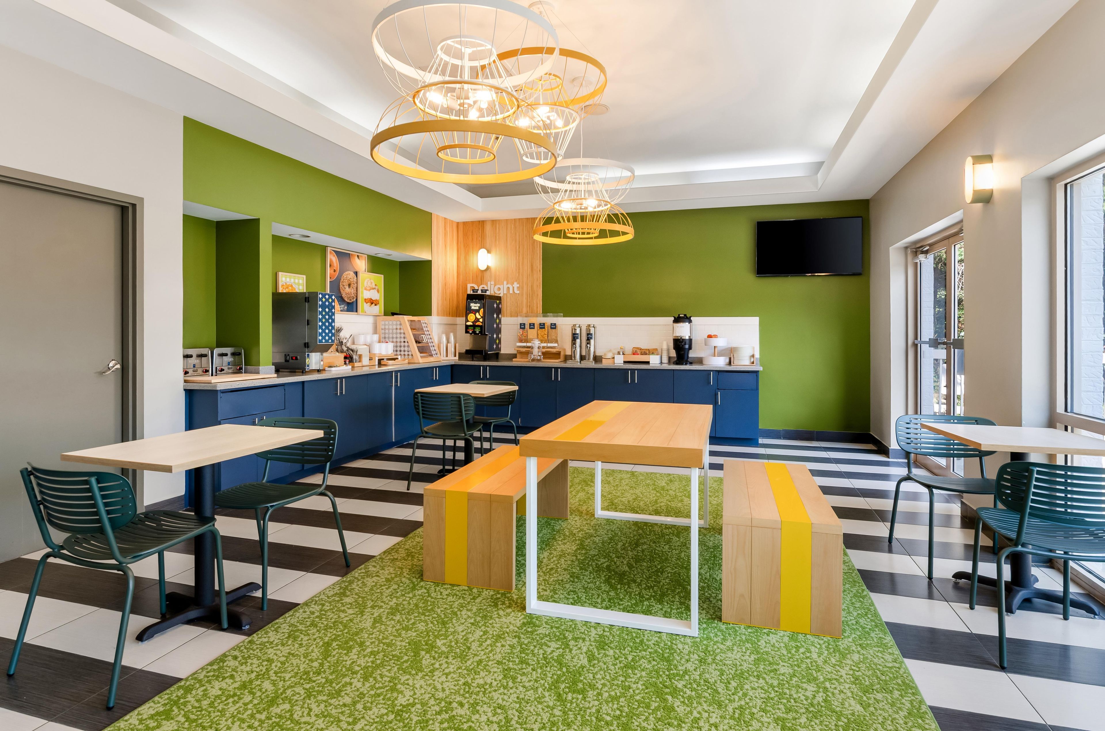 Um café moderno com piso xadrez, paredes verdes e azuis, e área de estar central com mesas e cadeiras.