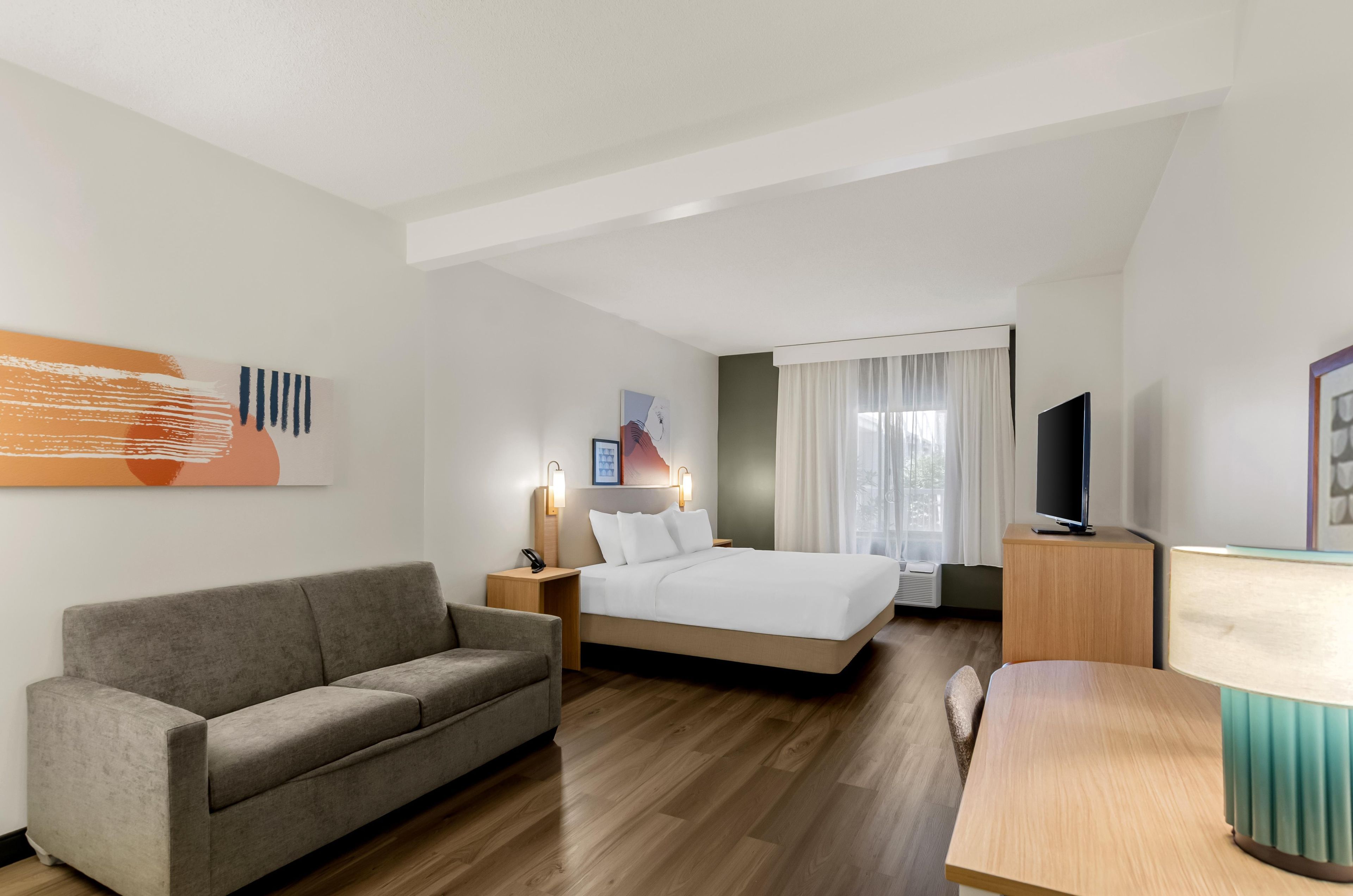 A hotel room with a bed, a sofa, a desk, a TV, and artwork on the walls.