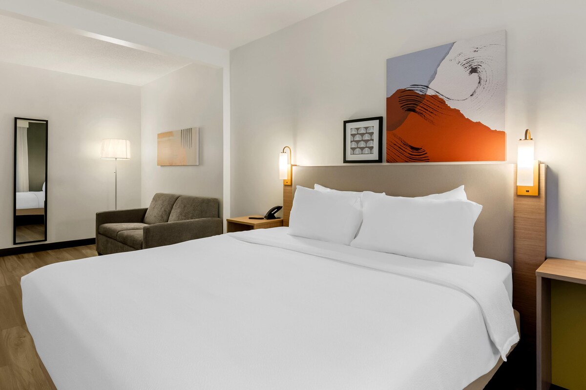 Una camera d'albergo con un letto grande, una sedia, un comodino con una lampada, uno specchio e opere d'arte alle pareti.