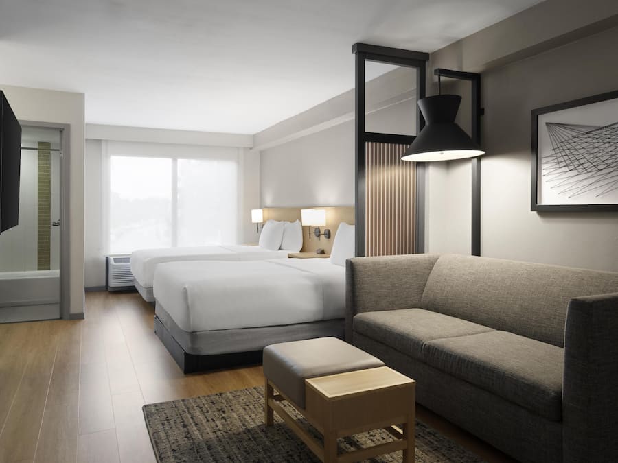 A modern hotel room with two beds, a sofa, a small wooden table, and a large window.