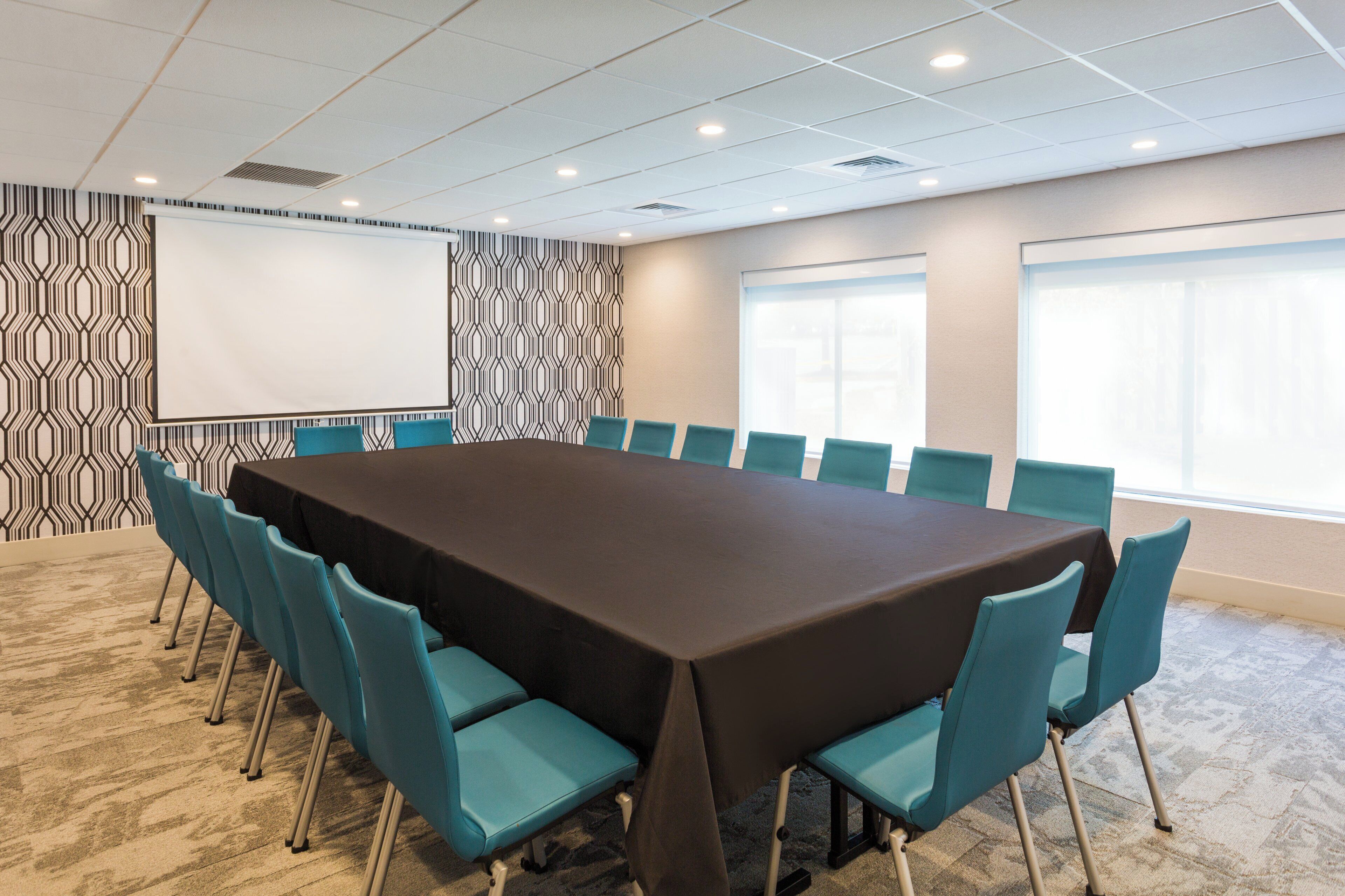 Un salón de conferencias con una larga mesa, sillas color aguamarina, una pantalla de proyector, paredes con diseño y ventanas amplias.