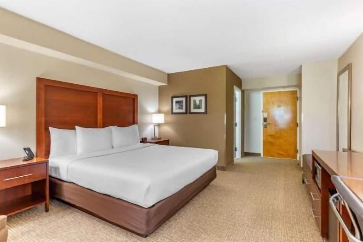 Quarto de hotel com cabeceira de madeira, uma cama grande, mesa de cabeceira com abajur, escrivaninha e guarda-roupa.