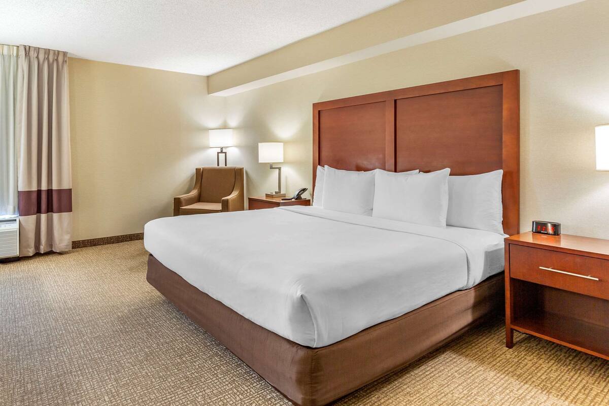 Quarto de hotel com uma cama grande, cabeceira de madeira, mesa de cabeceira com luminária e uma cadeira.