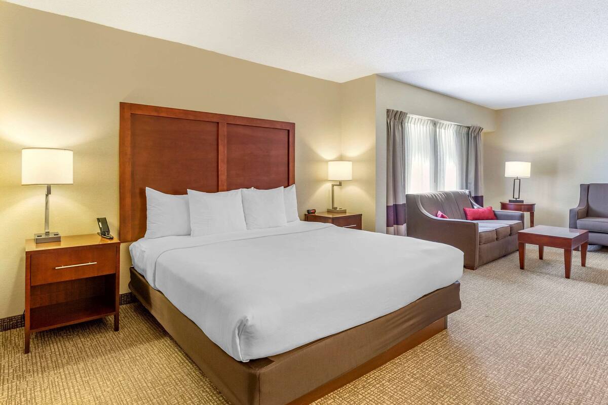 Quarto de hotel com uma cama grande, duas mesas de cabeceira com luminárias, um sofá, uma poltrona e uma pequena mesa.