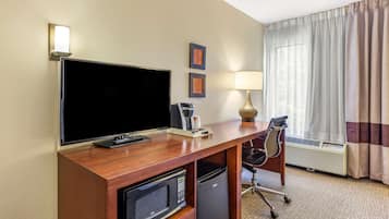 A hotel room with a wooden desk, a flat-screen TV, a microwave, a mini-fridge, a lamp, a chair, and a large window with curtains.