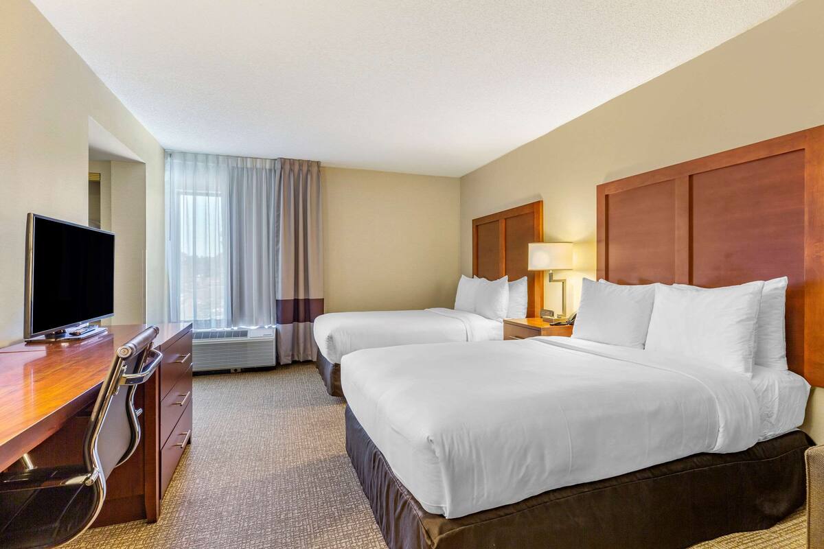 Quarto de hotel com duas camas, uma mesa de trabalho com televisão, uma luminária e uma janela com cortinas.