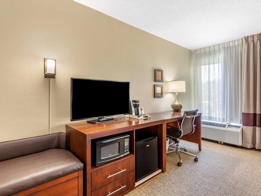 A hotel room with a wooden desk, a flat-screen TV, a microwave, a mini-fridge, a leather sofa, a desk lamp, and a large window with curtains.
