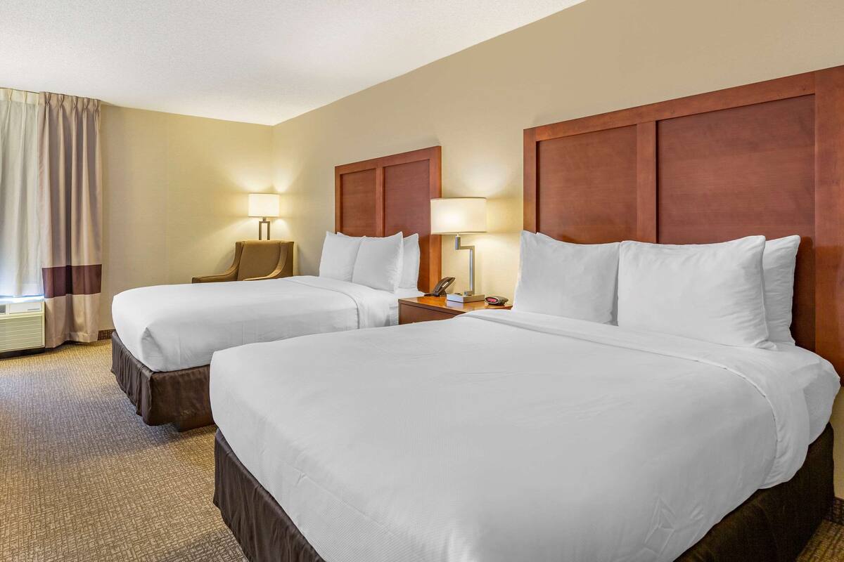Quarto de hotel com duas camas, cabeceira de madeira, luminárias de cabeceira, mesa de cabeceira e janela com cortinas.
