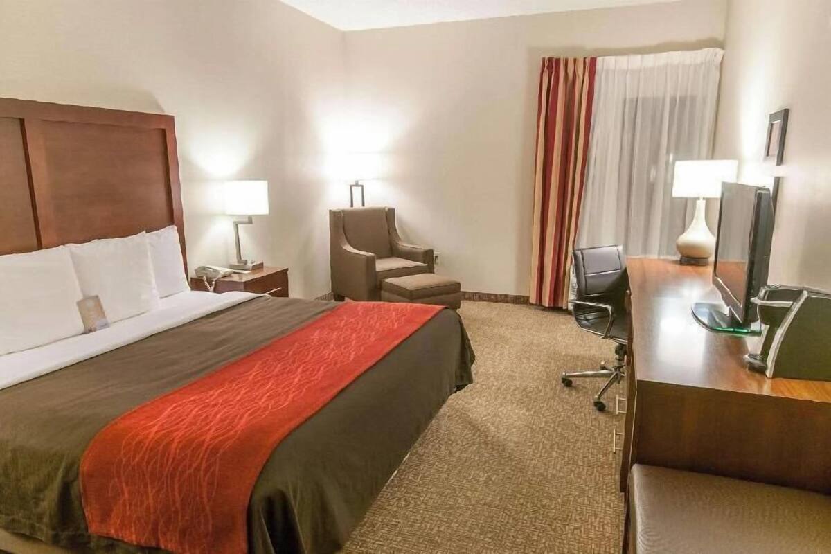 Quarto de hotel com cama, escrivaninha, cadeira, luminária e janela com cortinas.