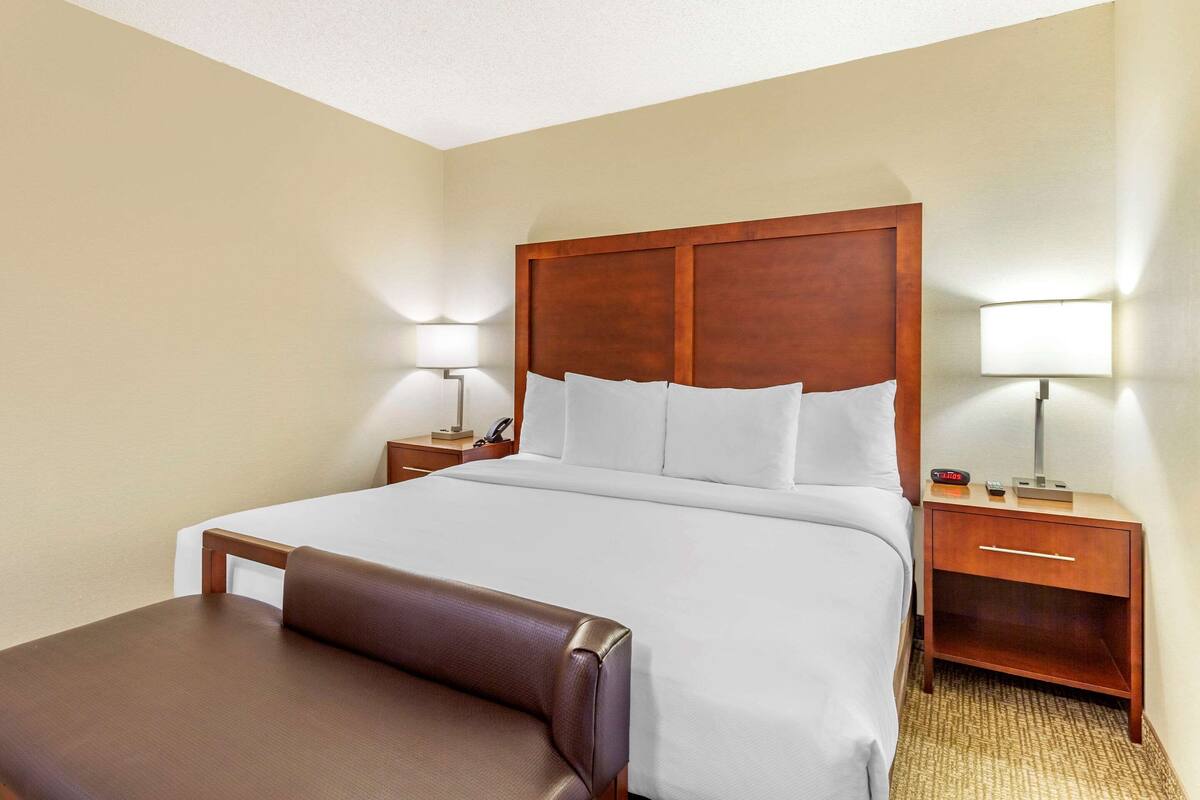 Quarto de hotel com uma cama grande, cabeceira de madeira, mesa de cabeceira com abajur e pequena escrivaninha com telefone e relógio.