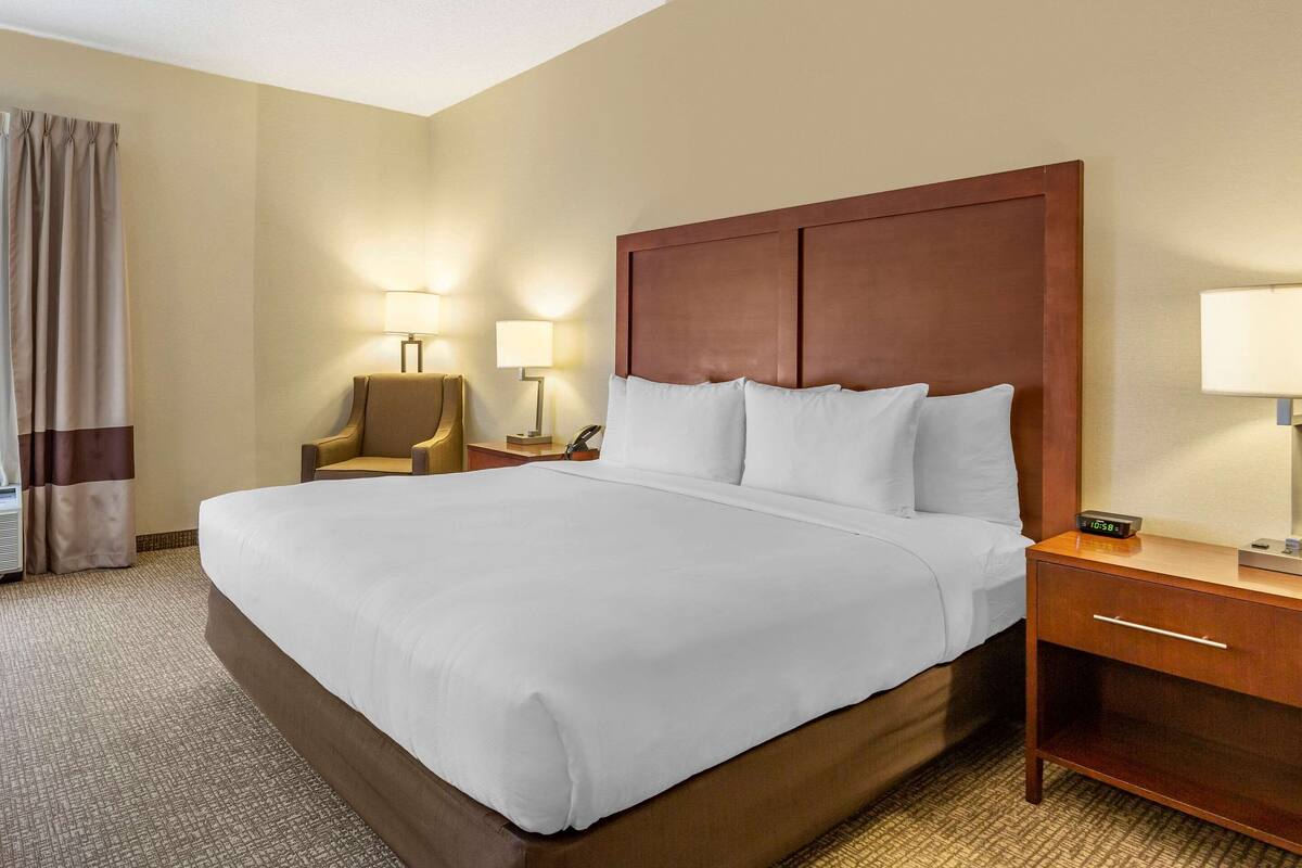 Quarto de hotel com cama grande, cabeceira de madeira, duas mesas de cabeceira com luminárias, uma cadeira e uma cortina.