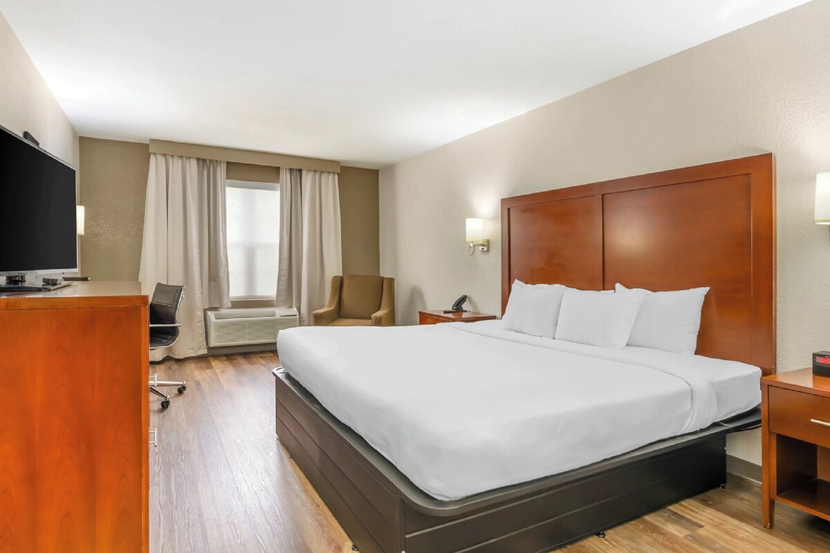 A hotel room with a large bed, a desk with a flat-screen TV, a chair, a desk lamp, and a window with curtains.