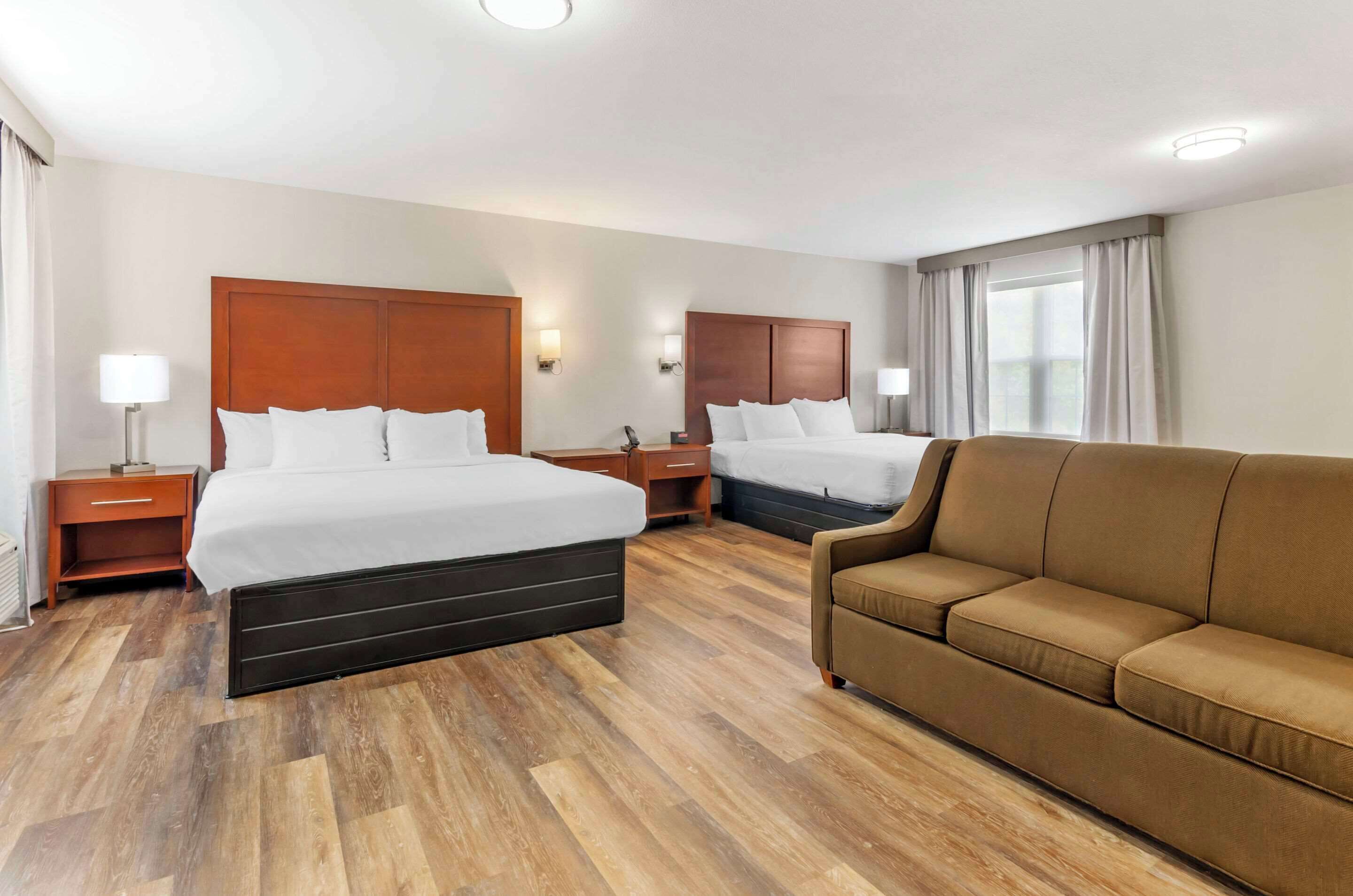 A hotel room with two beds, a brown sofa, wooden flooring, and a window with curtains.