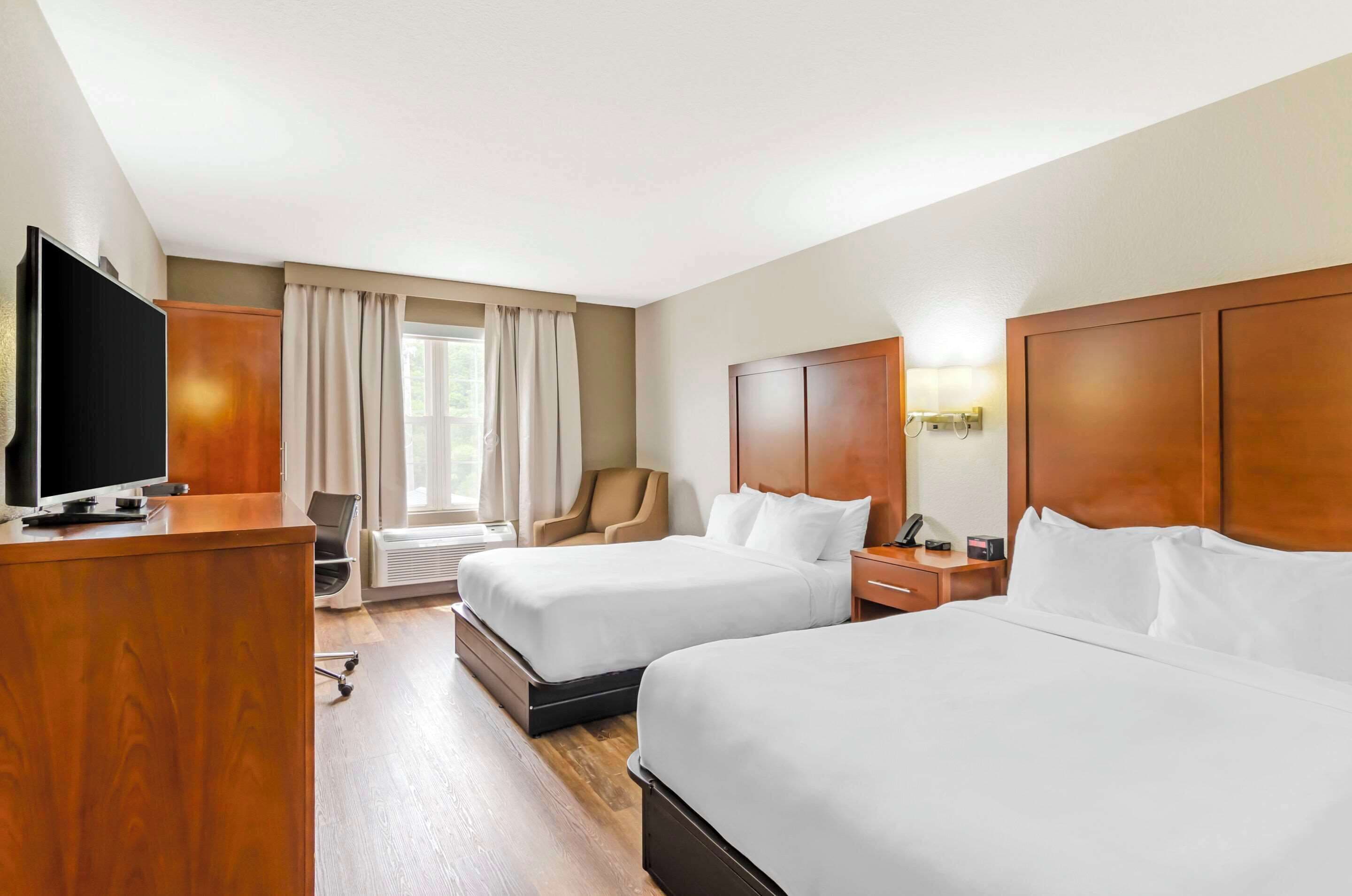 Een hotelkamer met twee bedden, een bureau met een flatscreen televisie, een stoel, een raam met gordijnen en een nachtkastje met een lamp.