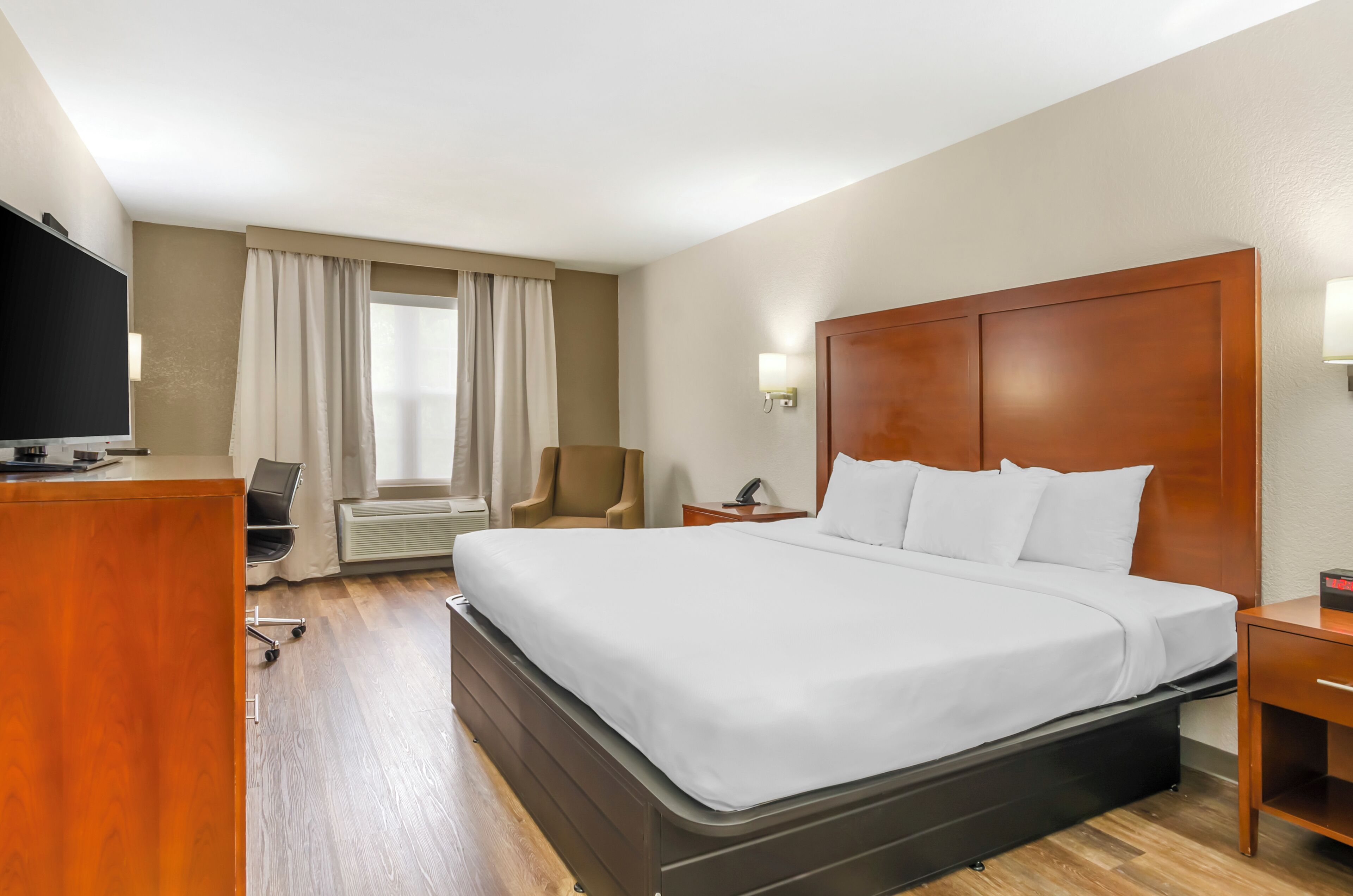 A hotel room with a large bed, a desk with a flat-screen TV, a chair, an air conditioning unit, and a window with curtains.