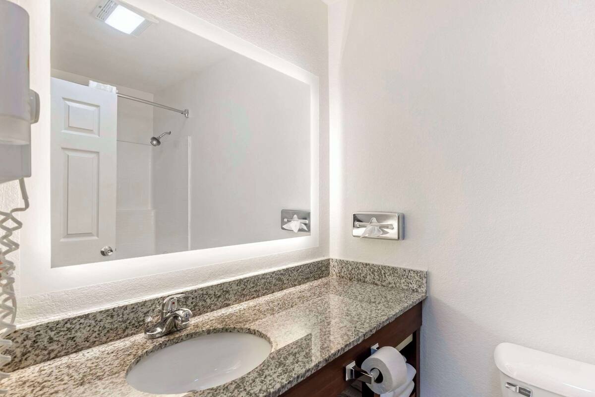 A bathroom with a granite countertop sink, a large mirror, a towel dispenser, and a toilet.