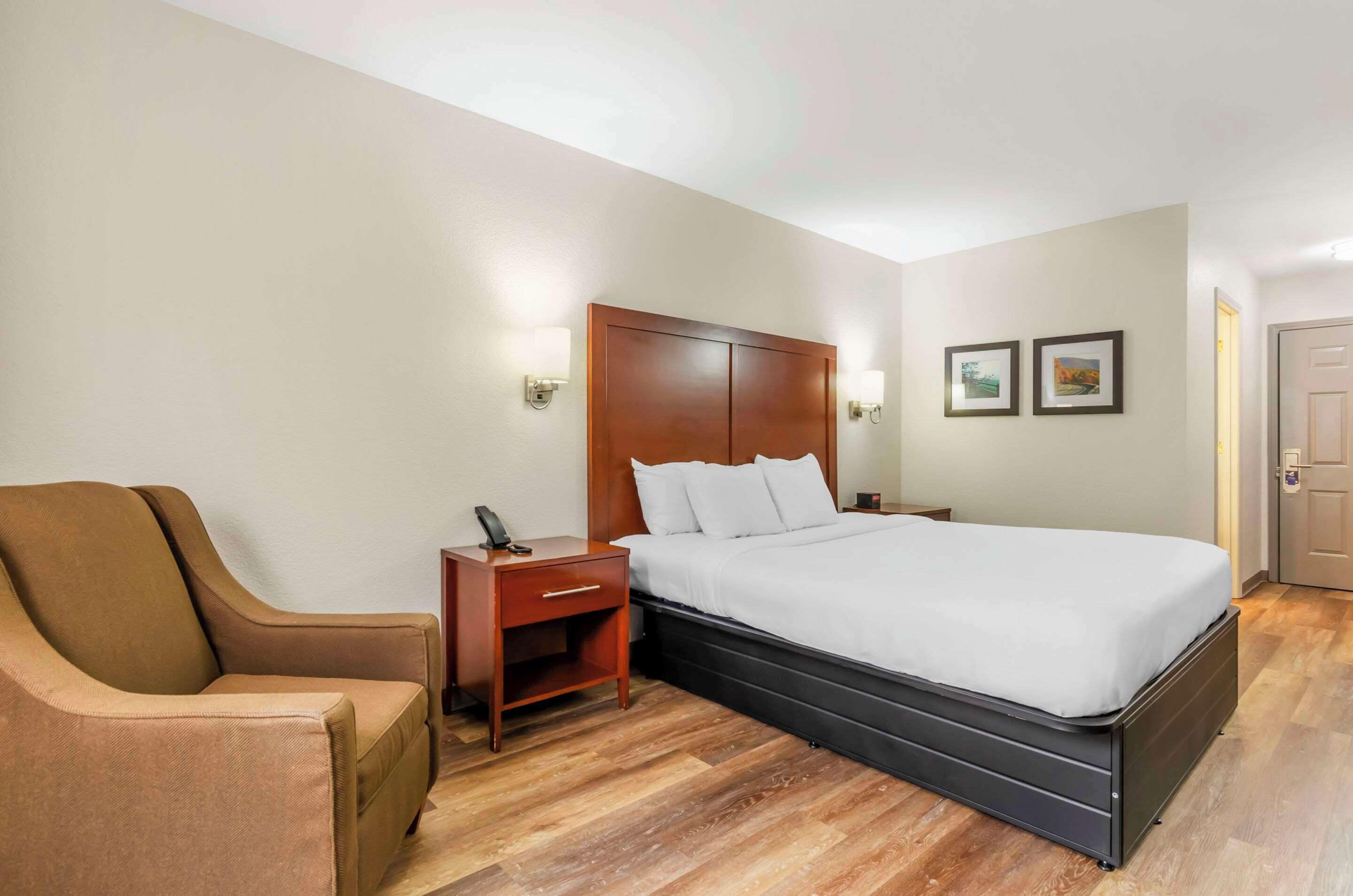 A hotel room with a bed, a wooden nightstand, a brown armchair, and two wall-mounted lamps.