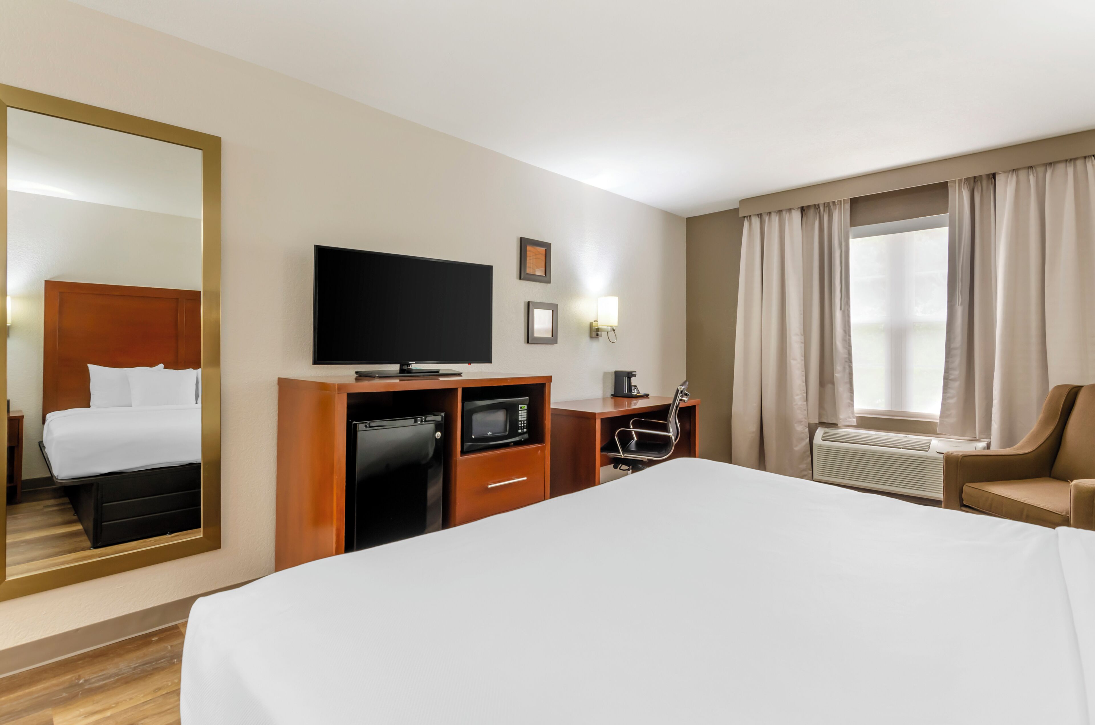 Ein Hotelzimmer mit einem großen Bett, einem Flachbildfernseher, einem Schreibtisch mit Mikrowelle, einem Sessel und einem Fenster mit Vorhängen.