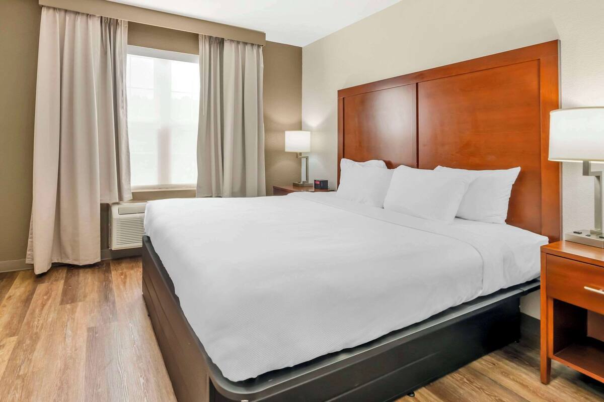 A hotel room with a large bed, wooden headboard, a nightstand with a lamp, and a window with curtains.