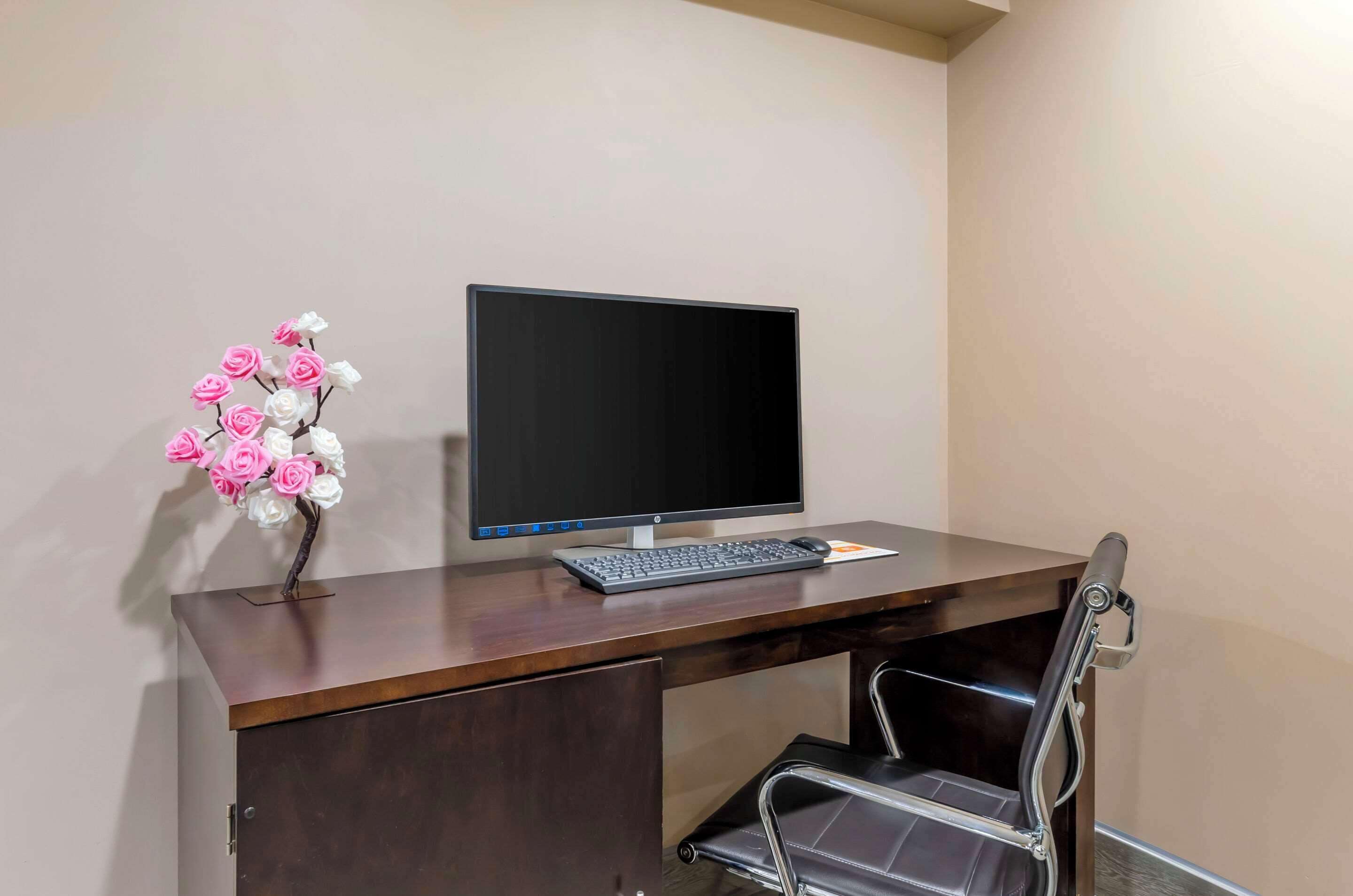 A wooden desk with a computer monitor, keyboard, and a vase with pink and white flowers.