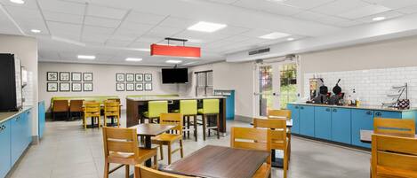 A modern restaurant with wooden tables and chairs, a kitchen area, and framed pictures on the walls.