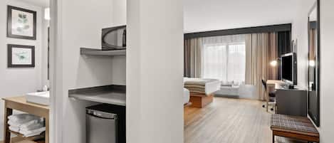A modern hotel room with a kitchenette, a bed with a view, a desk, and a television.