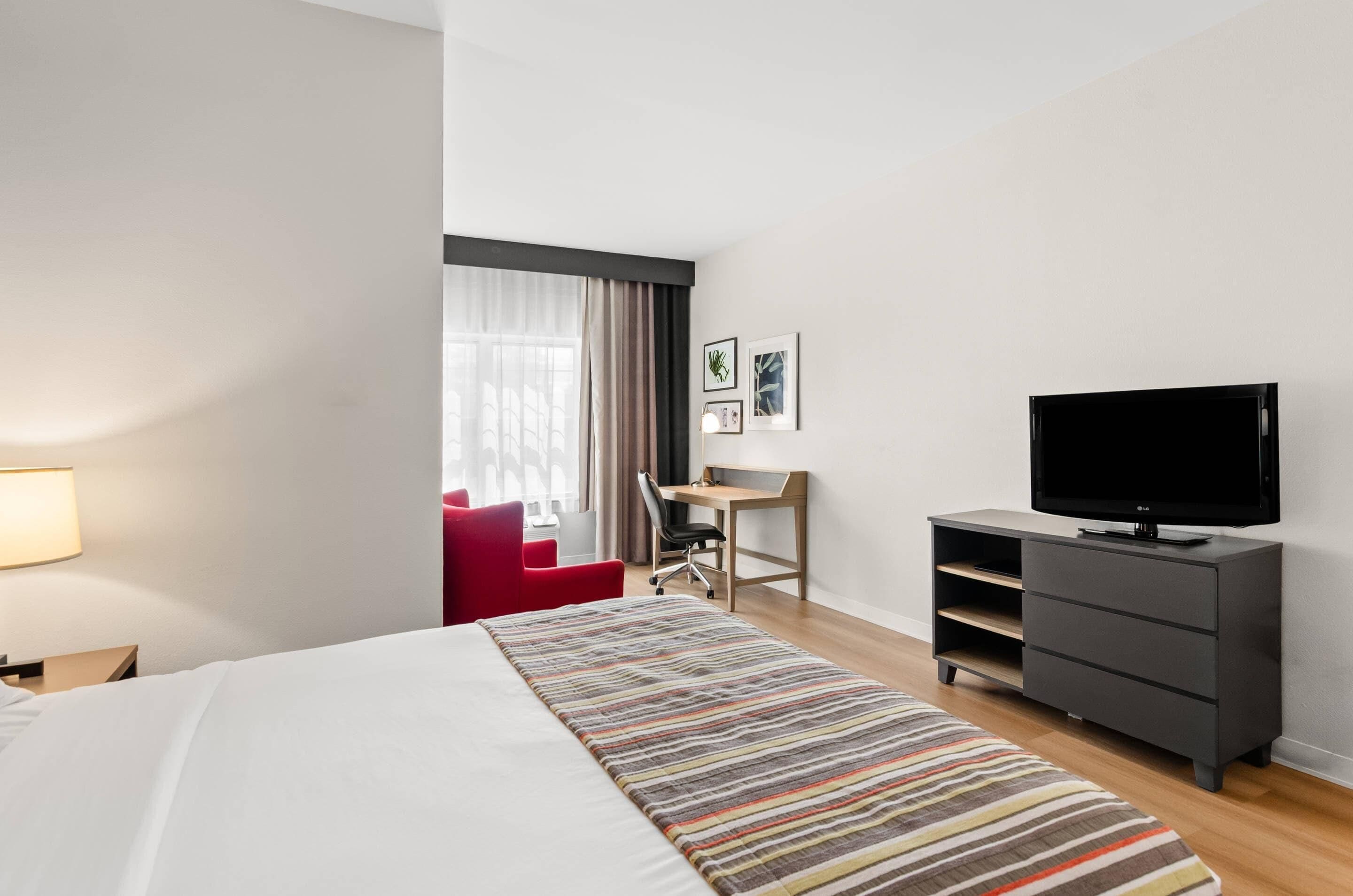 A hotel room with a bed, a desk, a chair, a television, and a red armchair.