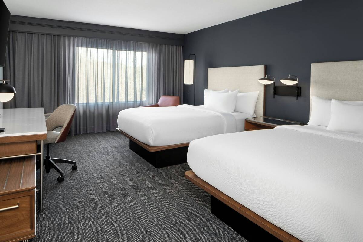 Habitación de hotel con dos camas, un escritorio, una silla y un ventanal con cortinas.