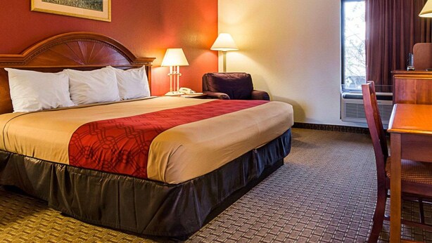 Quarto de hotel com uma cama grande, duas luminárias de cabeceira, uma cadeira, uma escrivaninha e uma janela com cortinas.