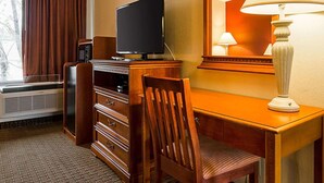 Quarto de hotel com televisão de tela plana sobre uma cômoda de madeira, uma cadeira, uma escrivaninha com luminária e um espelho grande.