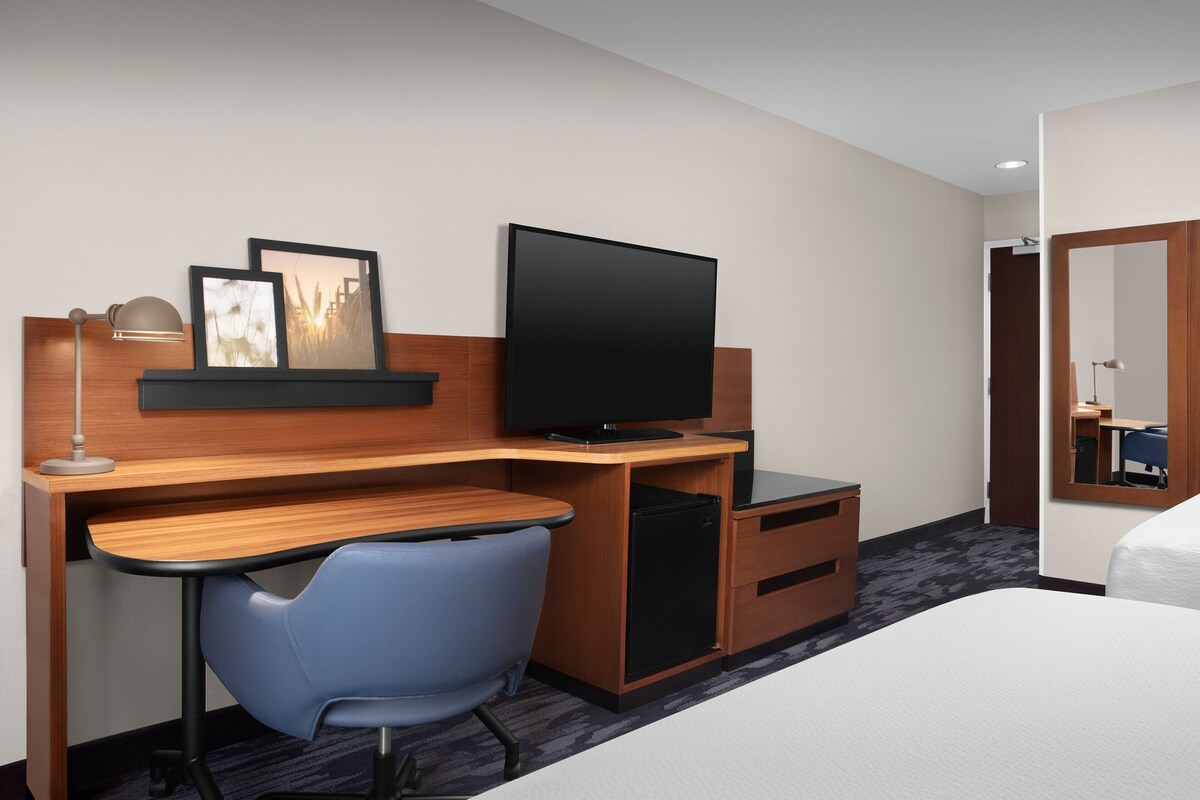 A hotel room with a wooden desk, a flat-screen TV, a blue chair, a bed, and a mirror.
