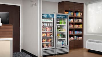 A corner of a hotel room with a refrigerator stocked with various food items, a small shelf with snacks, and a large window with blinds.