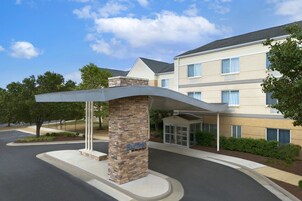 Entrada de hotel con un pilar de piedra que indica "Fairfield by Marriott", una galería techada y un estacionamiento.