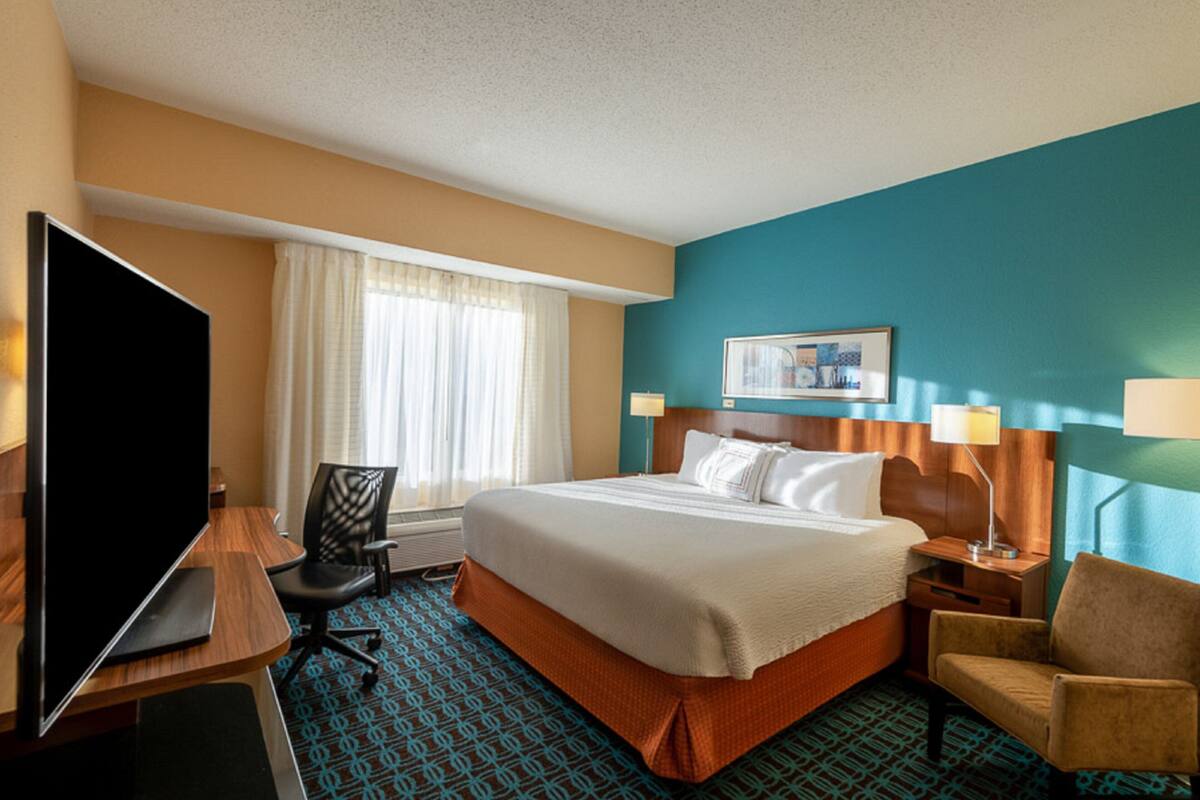 A hotel room with a bed, a desk with a TV, a chair, a window with curtains, and a painting on the wall.