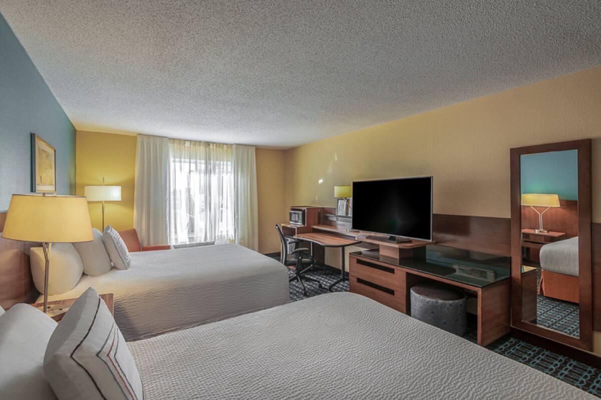 A hotel room with two beds, a desk with a flat-screen TV, a chair, a lamp, and a mirror.