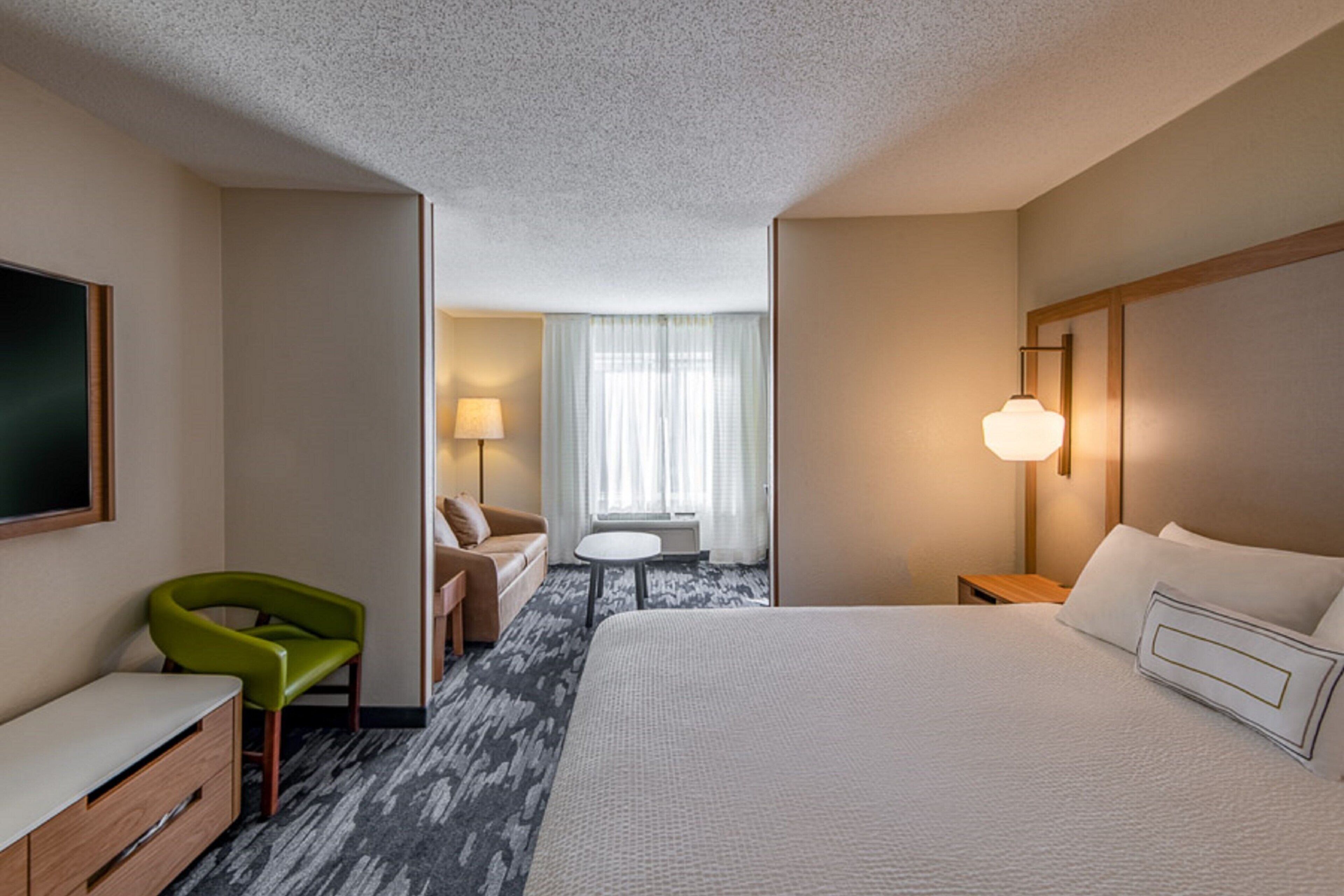 Ein Hotelzimmer mit einem großen Bett, einem grünen Sessel, einem Schreibtisch, einer Lampe und einem Fenster mit Vorhängen.