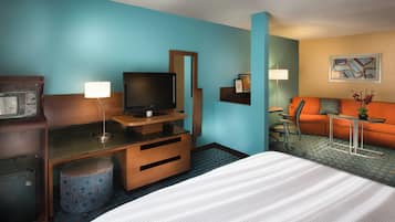 A hotel room with a bed, a desk with a television, a sofa, and a small table with a lamp.
