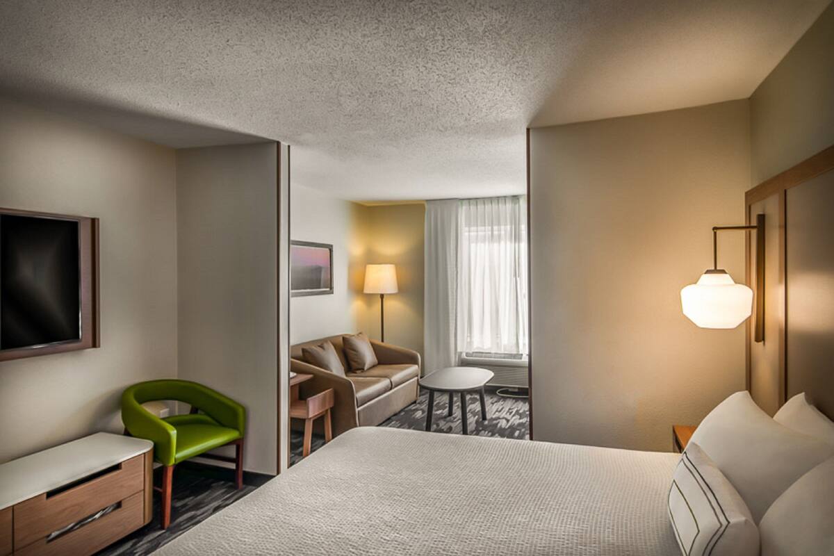 A hotel room with a large bed, a green chair, a sofa, a small table, a lamp, and a painting on the wall.
